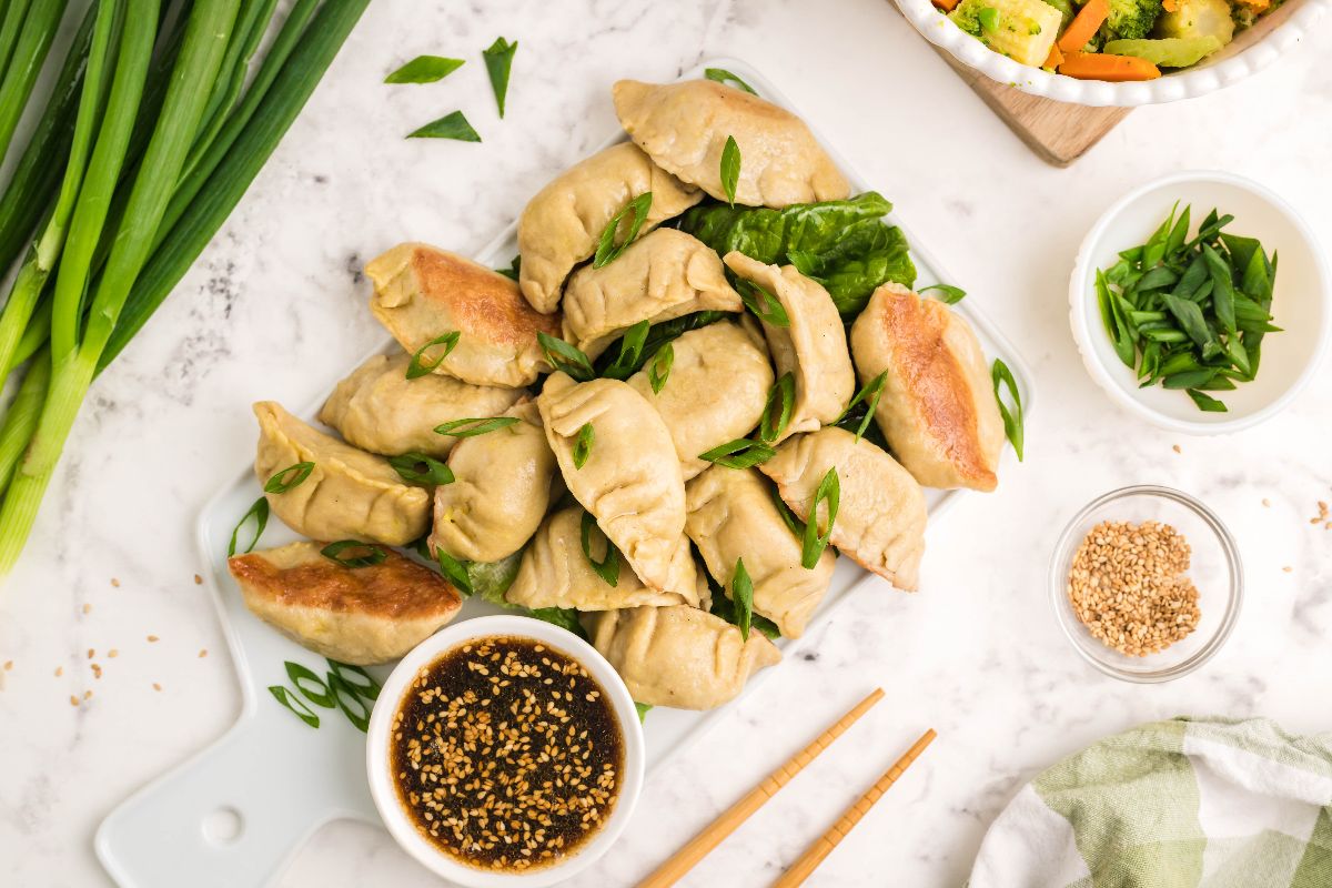 A plate of gluten free potstickers garnished with green onions, served with dipping sauce, sesame seeds, and vegetables.