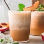 Two frothy peach smoothies in glasses, garnished with peach slices and mint, evoke the refreshing vibe of a Peach Bellini cocktail recipe, with ice and peaches nearby.