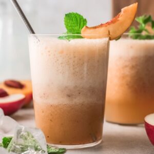 Two frothy peach smoothies in glasses, garnished with peach slices and mint, evoke the refreshing vibe of a Peach Bellini cocktail recipe, with ice and peaches nearby.