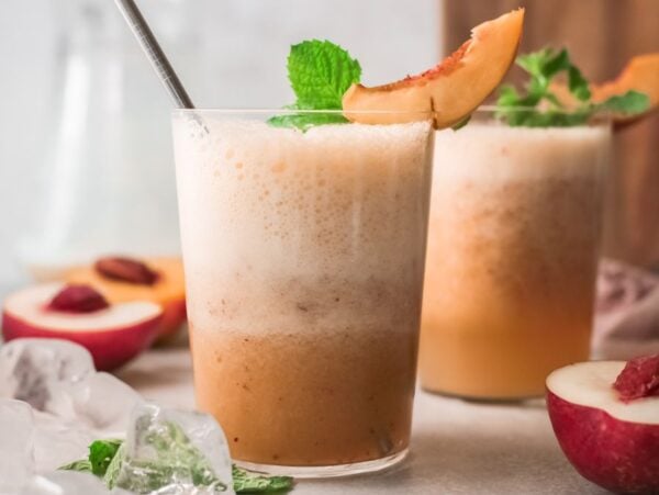 Two frothy peach smoothies in glasses, garnished with peach slices and mint, evoke the refreshing vibe of a Peach Bellini cocktail recipe, with ice and peaches nearby.