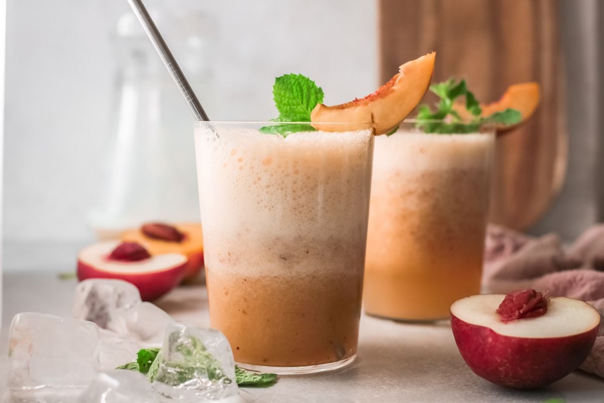 Two frothy peach smoothies in glasses, garnished with peach slices and mint, evoke the refreshing taste of a classic peach bellini. Enjoy this recipe with peach juice, served over ice with fresh peaches nearby.