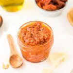 Small jar of red pesto with a wooden spoon, surrounded by pine nuts, parmesan, and sun-dried tomatoes—an easy pasta sauce and perfect red pesto recipe for any dish.