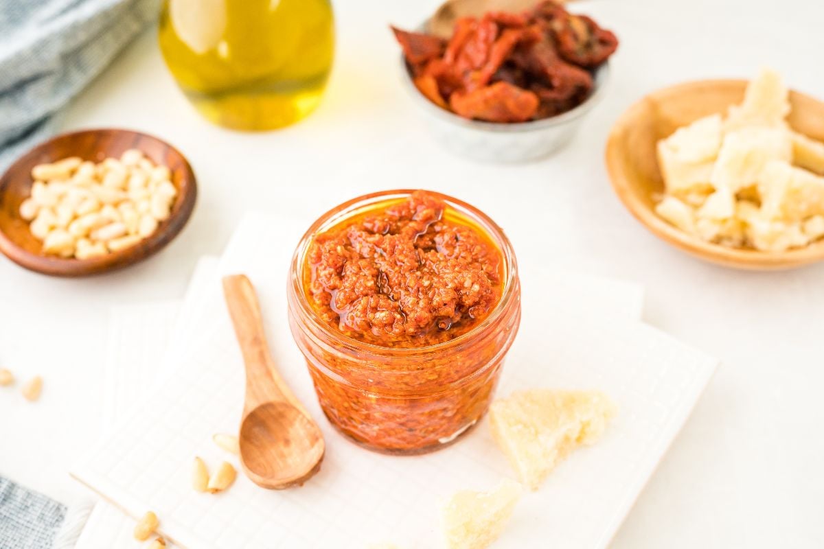 A jar of sun dried tomato pesto with a wooden spoon, surrounded by cheese, pine nuts, sun-dried tomatoes, and olive oil—an easy pasta sauce for any red pesto recipe.