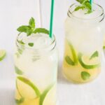 Two mason jars of a refreshing tropical mocktail with lime slices, mint leaves, ice, and green straws sit on a light wooden table.
