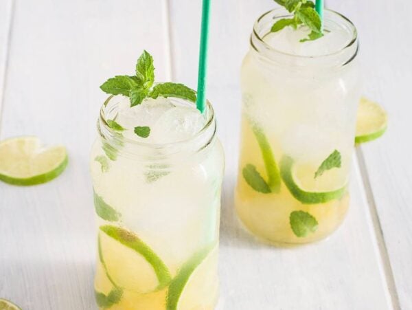 Two mason jars of a refreshing tropical mocktail with lime slices, mint leaves, ice, and green straws sit on a light wooden table.