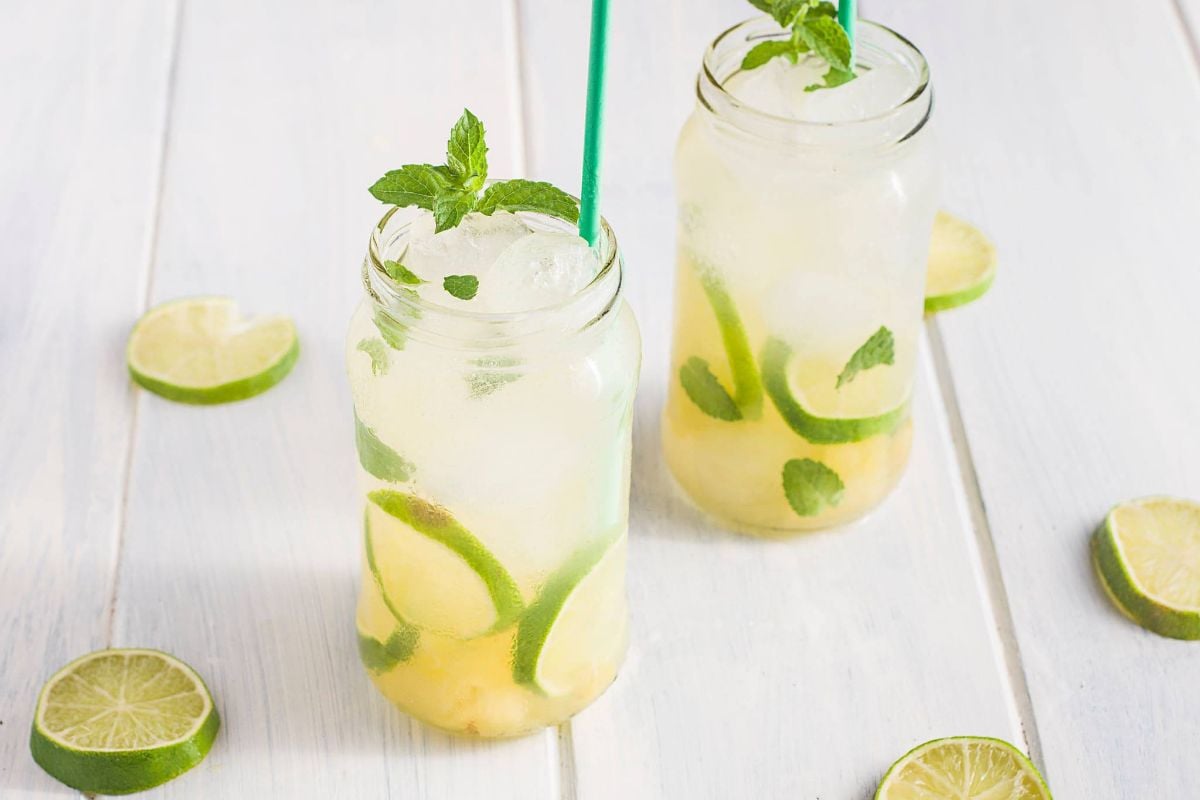 Two mason jars of lime and mint lemonade with straws, surrounded by lime slices on a white wooden surface—perfect inspiration for a refreshing tropical mocktail.