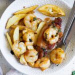 A plate with shrimp, potato wedges, grilled chicken, and a lemon wedge captures the spirit of surf and turf dining. A fork and knife rest on the side, ready for you to enjoy this delicious combination.