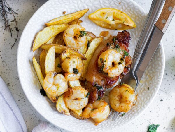 A plate with shrimp, potato wedges, grilled chicken, and a lemon wedge captures the spirit of surf and turf dining. A fork and knife rest on the side, ready for you to enjoy this delicious combination.