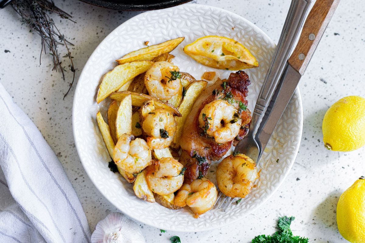 White bowl with butter herbed shrimp, potato wedges, chicken, a lemon wedge, and a fork and knife on a light countertop.