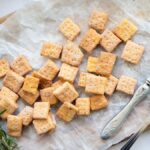 Square rosemary cheddar cheese crackers on parchment paper with two forks and sprigs of rosemary, beautifully arranged on a wooden board—a perfect homemade cracker recipe for any occasion.