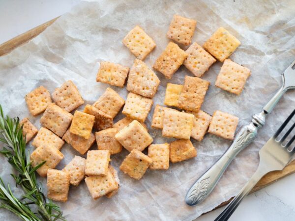 Square rosemary cheddar cheese crackers on parchment paper with two forks and sprigs of rosemary, beautifully arranged on a wooden board—a perfect homemade cracker recipe for any occasion.
