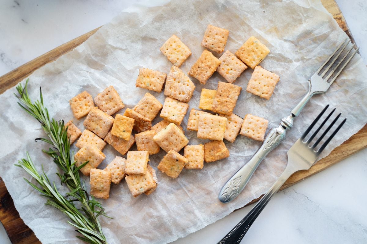 Rosemary and cheddar crackers on parchment paper with rosemary sprigs and two forks rest on a wooden board.