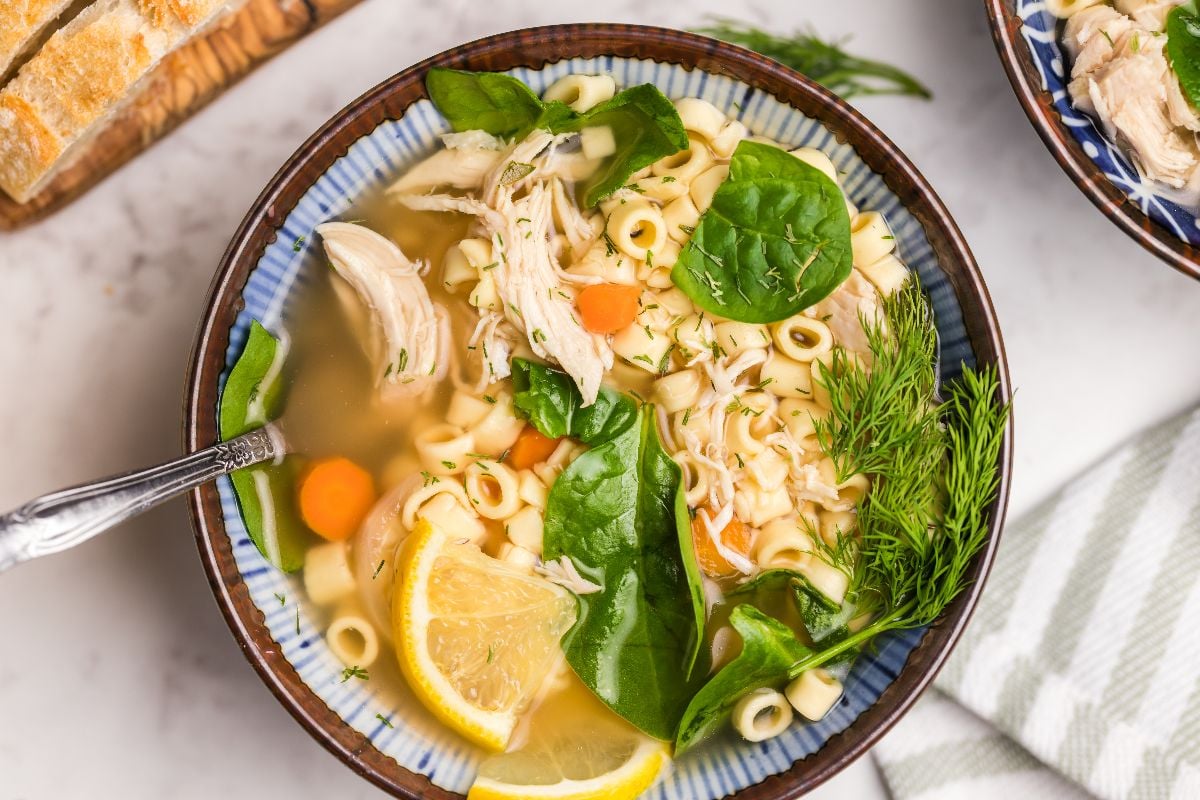 A bowl of Greek lemon chicken soup with spinach, carrots, dill, and lemon slices, served with bread on the side.