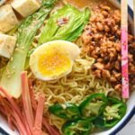 Bowl of ramen with egg, tofu, bok choy, ham, jalapeños, minced meat, and chopsticks on the side—perfect for fans of Spicy Chicken Miso Ramen or anyone craving ramen with chicken and tofu.