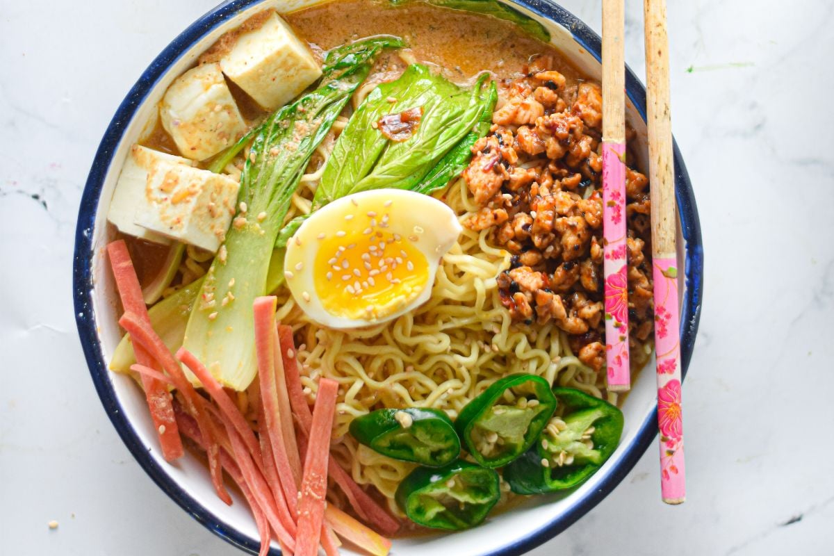 Bowl of ramen with noodles, bok choy, egg, tofu, sliced jalapeños, ham, and chopsticks—perfect for trying your favorite ramen recipe at home.