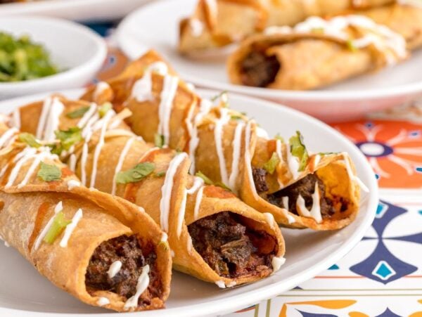 Three beef taquitos topped with sour cream and herbs on a white plate, with more taquitos in the background—an easy air fryer taquitos dish perfect for a gluten free Mexican dinner.