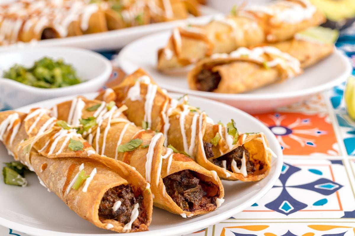 Three crispy beef taquitos made with an easy taquito recipe, topped with crema and cilantro on a white plate, with more taquitos in the background.
