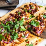 Rectangular BBQ Chicken Flatbread Pizza topped with cilantro, red onions, chicken, and cheese on a wooden board—one slice being served. Make flatbread pizza at home for a tasty barbecue chicken flatbread like Panera.