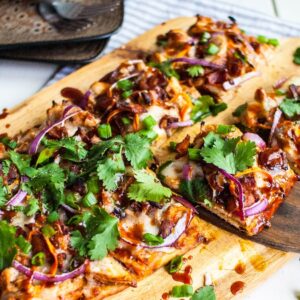 Rectangular BBQ Chicken Flatbread Pizza topped with cilantro, red onions, chicken, and cheese on a wooden board—one slice being served. Make flatbread pizza at home for a tasty barbecue chicken flatbread like Panera.