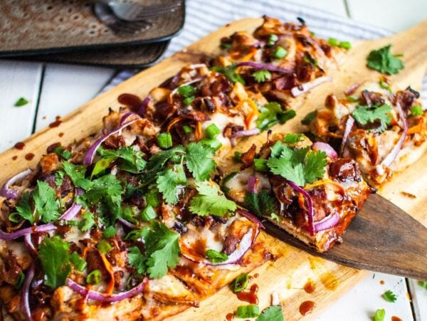 Rectangular BBQ Chicken Flatbread Pizza topped with cilantro, red onions, chicken, and cheese on a wooden board—one slice being served. Make flatbread pizza at home for a tasty barbecue chicken flatbread like Panera.