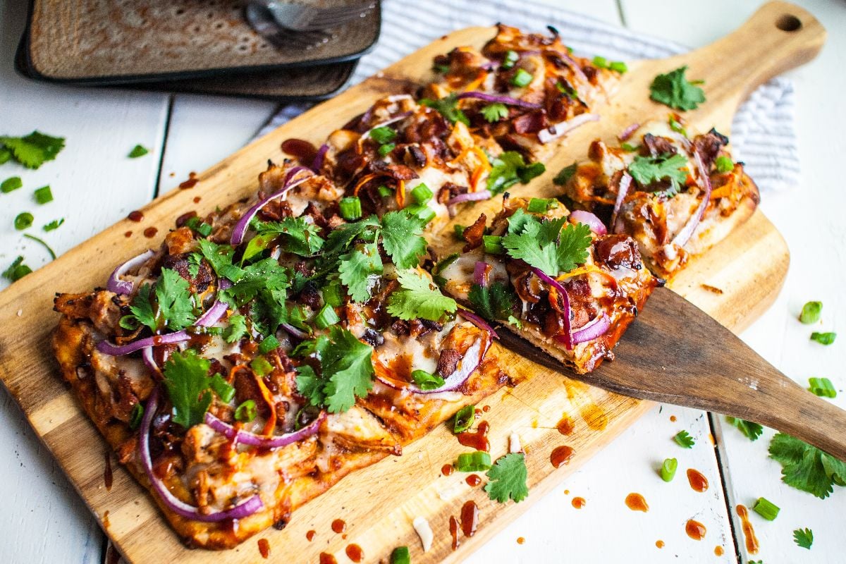 Rectangular pizza topped with red onions, cilantro, and sauce on a wooden board, one slice being served—enjoy easy BBQ chicken pizza inspired by Panera’s popular barbecue chicken flatbread right at home.