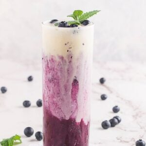 Tall glass of sparkling blueberry float, layered with cream and garnished with mint and blueberries, sits on a marble surface.