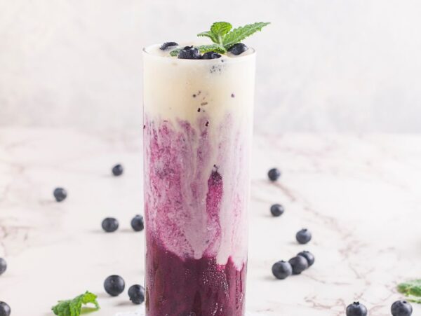 Tall glass of sparkling blueberry float, layered with cream and garnished with mint and blueberries, sits on a marble surface.