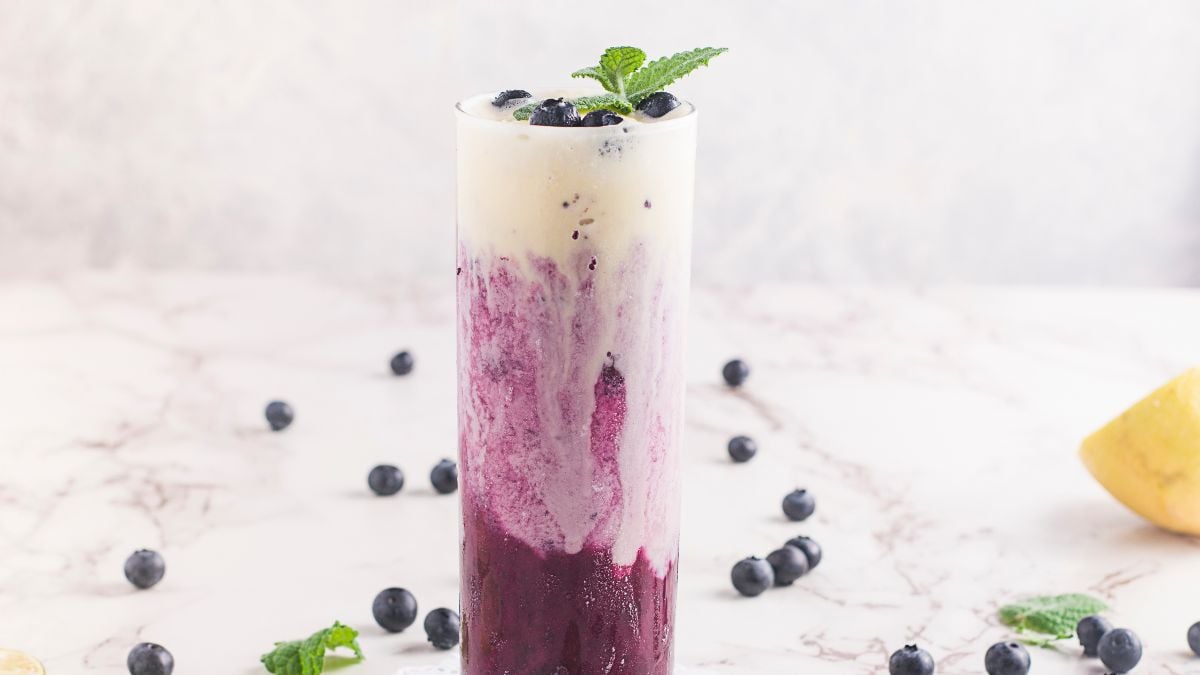 Tall glass of sparkling blueberry float, layered with cream and garnished with mint and blueberries, sits on a marble surface.