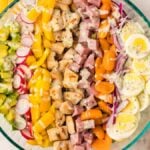 A glass bowl filled with a colorful chopped salad, topped with veggies, ham, eggs, and homemade ranch dressing for a filling dinner salad.