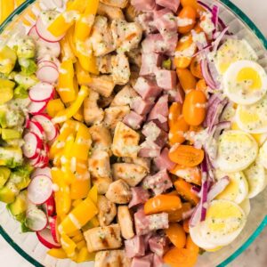 A glass bowl filled with a colorful chopped salad, topped with veggies, ham, eggs, and homemade ranch dressing for a filling dinner salad.