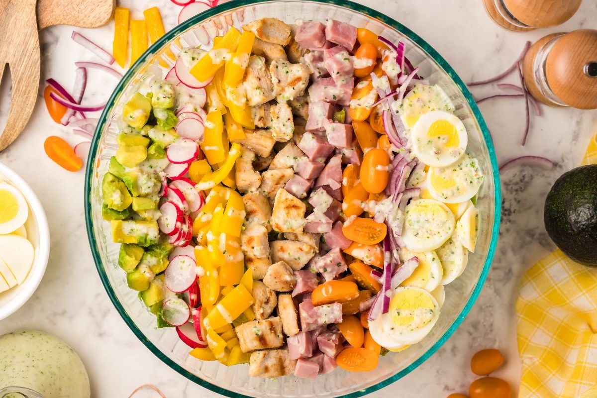 A colorful Cobb salad with eggs, ham, chicken, tomatoes, radishes, pickles, and yellow peppers in a glass bowl, served with homemade ranch dressing.