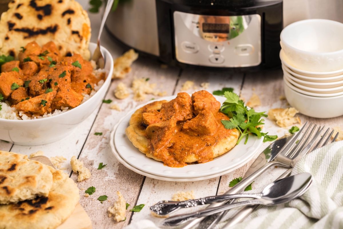 Plate of chicken tikka masala on fluffy naan bread, with rice, more naan, and a slow cooker in the background.