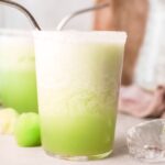 A frothy margarita with melon in a glass with a metal straw, surrounded by lime wedges and ice cubes—an easy tequila cocktail perfect as your new favorite summer cocktail recipe.