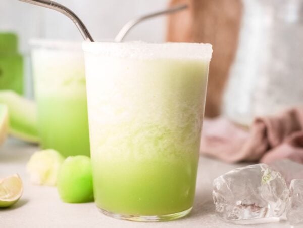 A frothy margarita with melon in a glass with a metal straw, surrounded by lime wedges and ice cubes—an easy tequila cocktail perfect as your new favorite summer cocktail recipe.