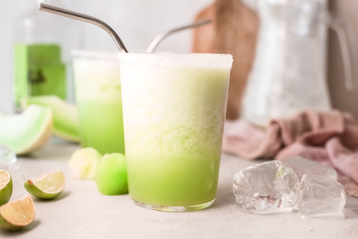 A glass of frothy honeydew cocktail with a metal straw, ice cubes, and lime wedges nearby—an easy tequila cocktail perfect for summer sipping.