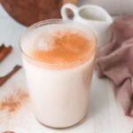 A glass of homemade horchata topped with cinnamon, with cinnamon sticks and a pink cloth nearby on a white surface.