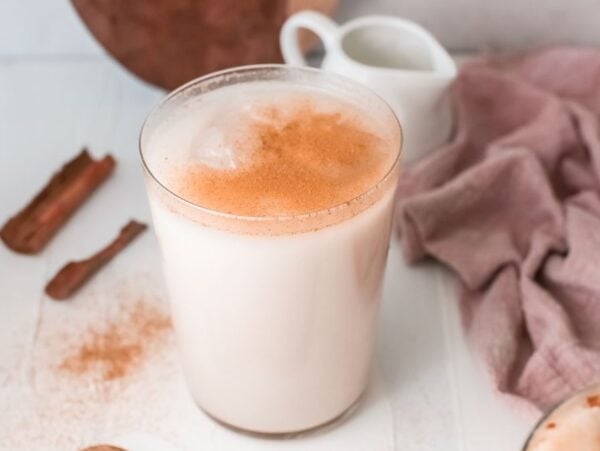 A glass of homemade horchata topped with cinnamon, with cinnamon sticks and a pink cloth nearby on a white surface.