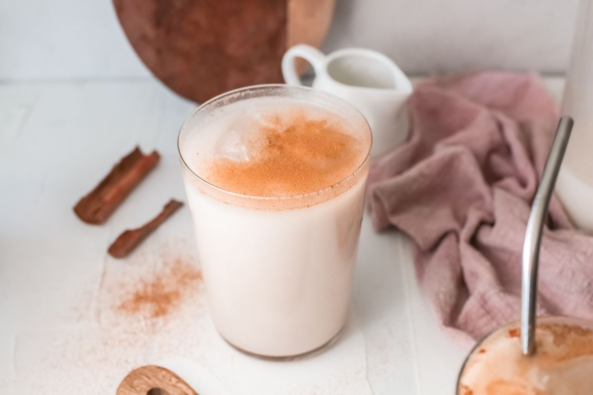 A glass of Mexican Horchata topped with cinnamon sits on a white table beside a cinnamon stick and a pink napkin, perfectly showcasing this classic iced rice milk beverage.