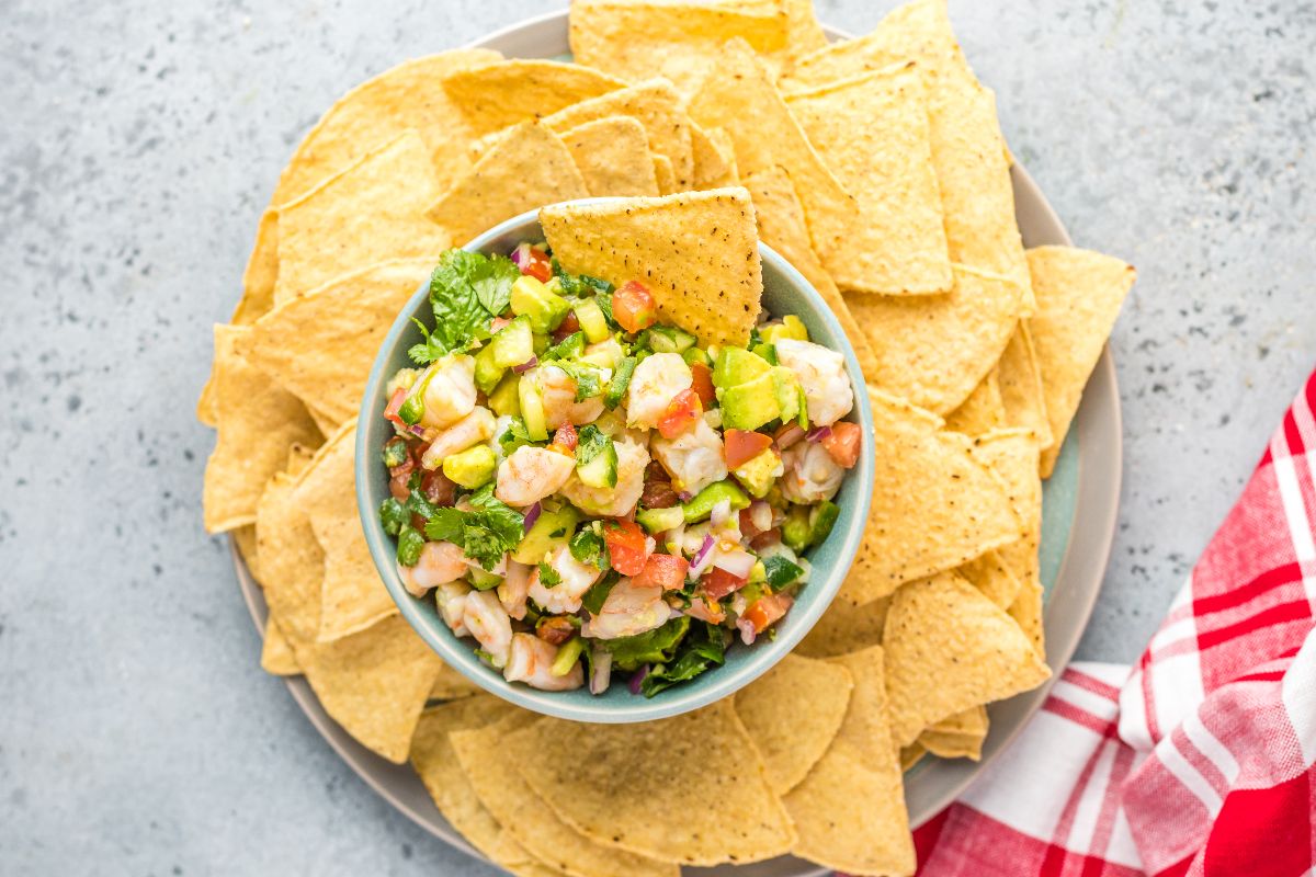 Bowl of Mexican ceviche with avocado, surrounded by tortilla chips on a plate, next to a red napkin—a delicious seafood ceviche recipe for any occasion.