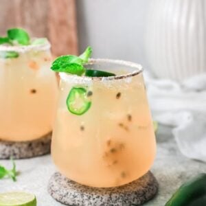 Two glasses of a light, iced passion fruit margarita garnished with mint and jalapeño slices on a stone coaster, with limes nearby—an easy margarita recipe perfect for summer refreshment.