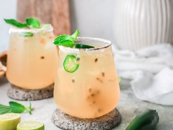 Two glasses of a light, iced passion fruit margarita garnished with mint and jalapeño slices on a stone coaster, with limes nearby—an easy margarita recipe perfect for summer refreshment.