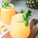 Two glasses of yellow tropical juice garnished with mint and lime on a wooden board, with pineapple in the background—a refreshing Summer drink with pineapple inspired by a classic Agua Fresca recipe.