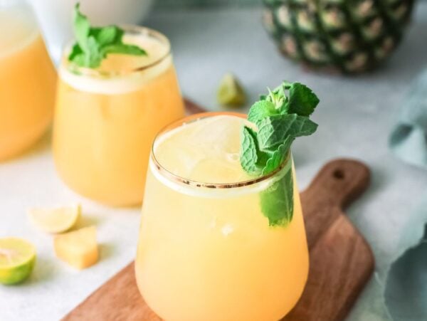 Two glasses of yellow tropical juice garnished with mint and lime on a wooden board, with pineapple in the background—a refreshing Summer drink with pineapple inspired by a classic Agua Fresca recipe.