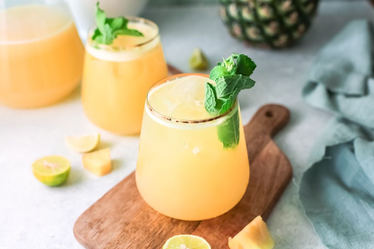 A glass of orange tropical drink with mint and lemon garnish on a wooden board, plus fruit pieces nearby—a refreshing drink recipe perfect for summer days.