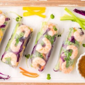 Four fresh Vietnamese spring rolls with colorful vegetables and shrimp on a tray, served with a spicy garlic sauce in a small bowl.