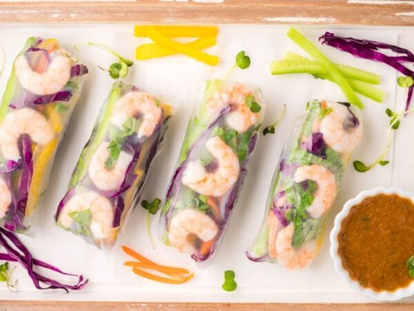 Four fresh Vietnamese spring rolls with colorful vegetables and shrimp on a tray, served with a spicy garlic sauce in a small bowl.
