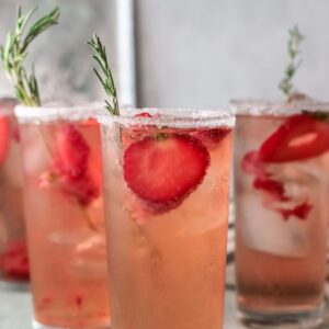 Four glasses of strawberry tequila cocktail with ice, garnished with rosemary and sugar rims, sit on a light table.