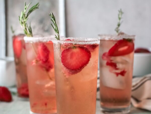 Four glasses of strawberry tequila cocktail with ice, garnished with rosemary and sugar rims, sit on a light table.