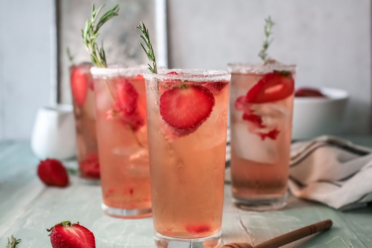 Three tall glasses of pink strawberry tequila cocktail with ice, garnished with rosemary and fresh strawberry slices.