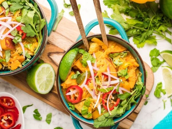Two bowls of colorful curry, inspired by a hearty Thai recipe with chicken, loaded with veggies, lime wedges, herbs, and sliced chili peppers on a marble surface.